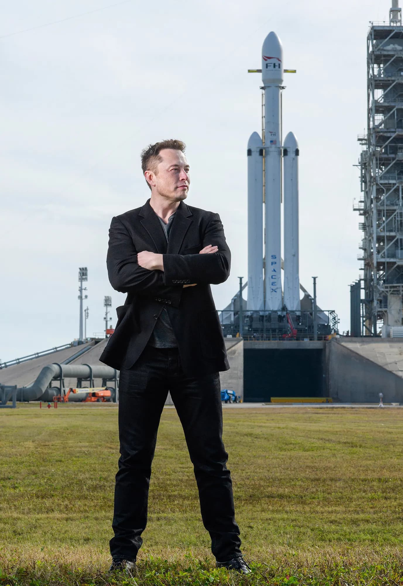 Elon Musk standing arms-crossed on a launch site lawn with a SpaceX Falcon Heavy rocket on the pad behind him