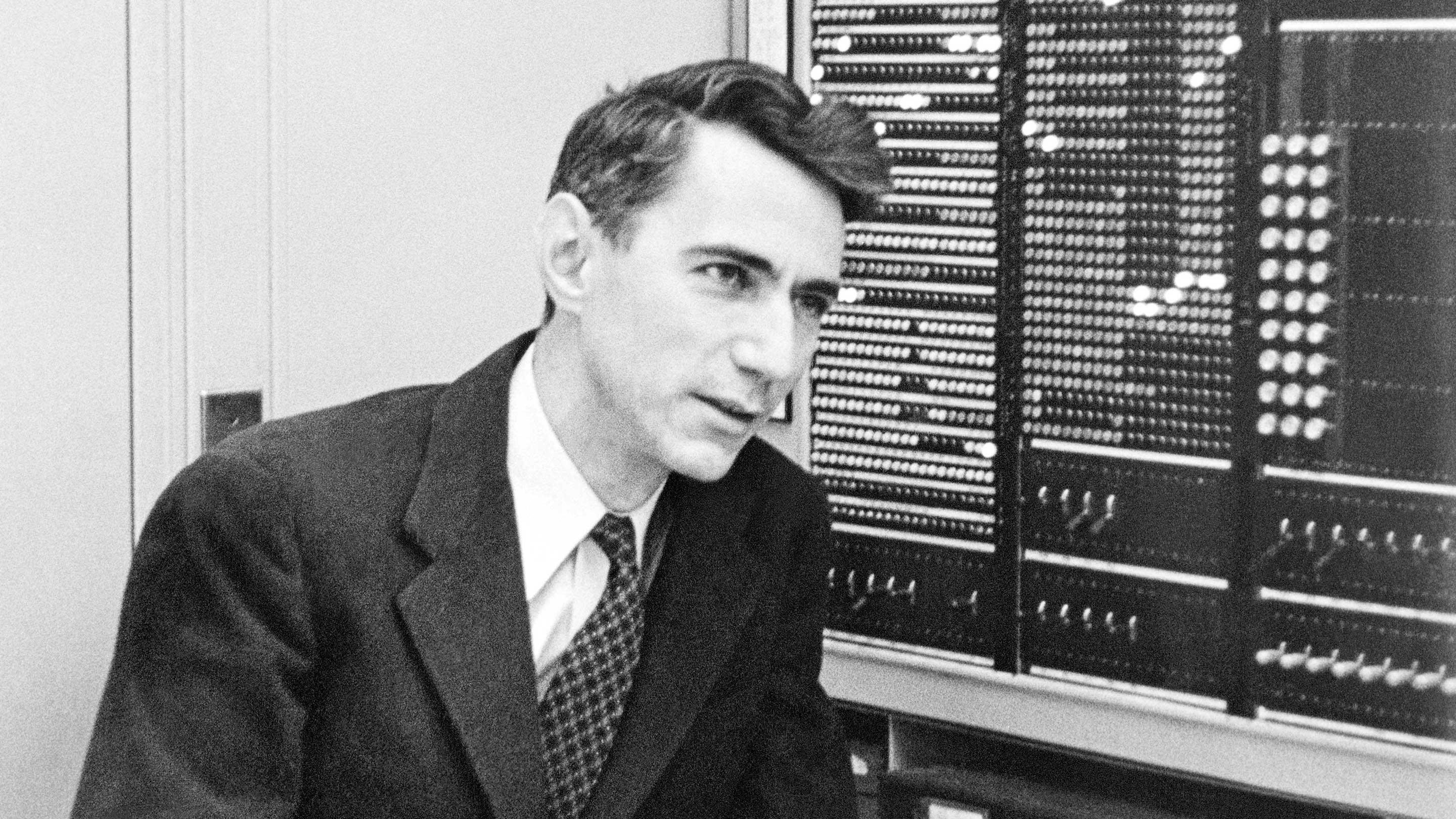Black-and-white photograph of Claude Shannon beside a wall of early computing relays at Bell Labs